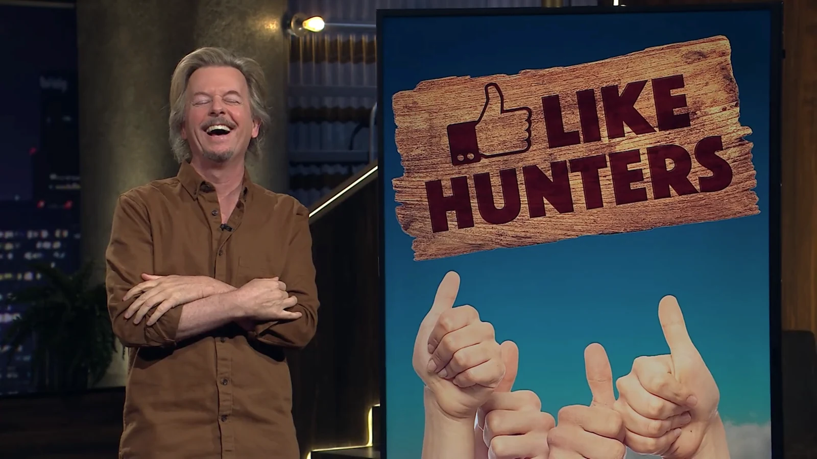 David Spade laughing beside Burt Rutherford’s Like Hunters monitor graphic
